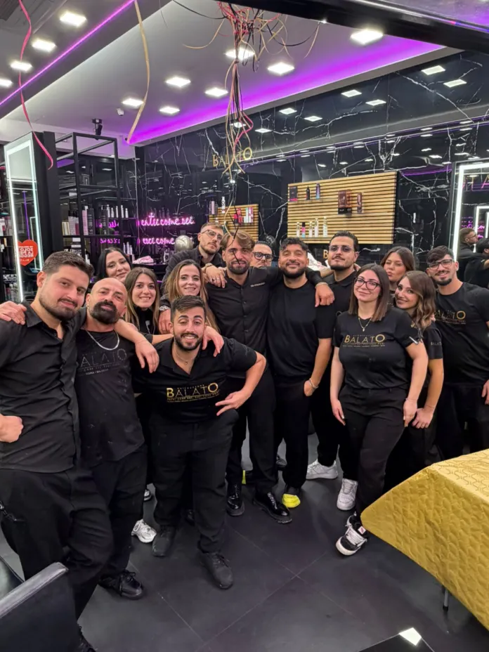 Immagine che mostra un gruppo di persone sorridenti all'interno di un salone di bellezza, celebrando il quinto anniversario del salone Balato con azioni di solidarietà.