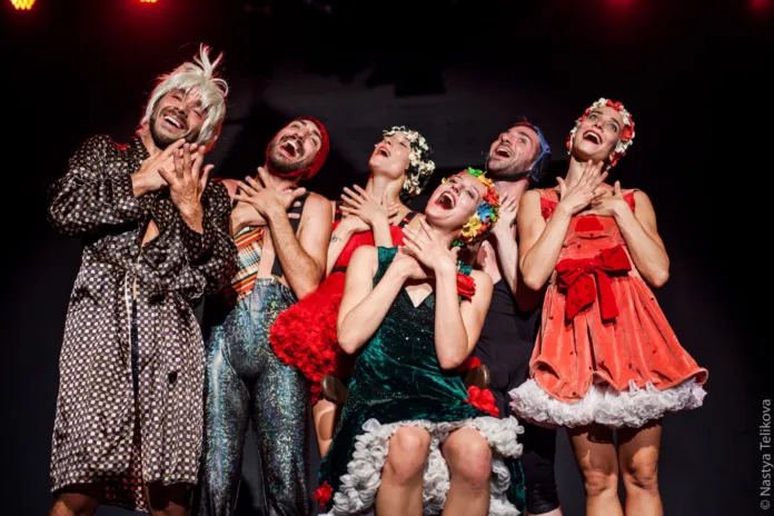 Sei interpreti in abiti colorati e di scena posano sul palco sotto luci rosse.