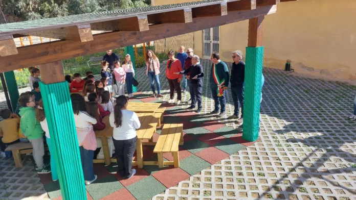 Persone riunite sotto una copertura in legno, accanto a tavoli e sedute colorate, in un’area esterna.