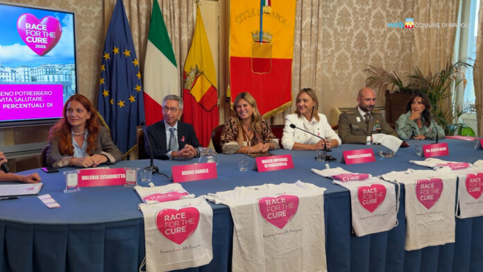 Conferenza stampa con diverse persone sedute al tavolo con bandiere e striscioni relativi alla Race for the Cure.