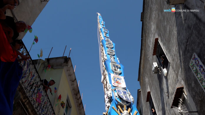 Giglio alto con decorazioni e scritte, visto dal basso tra due edifici durante la giornata.
