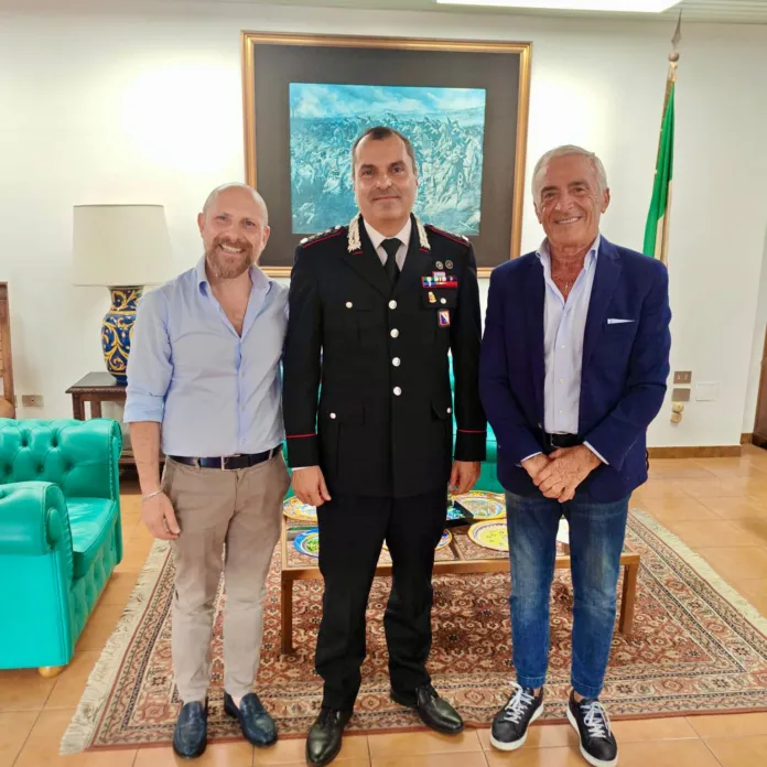 Fotografia di archivio che mostra tre uomini, tra cui un ufficiale dei Carabinieri in uniforme, durante un incontro istituzionale con i rappresentanti dei commercianti, in un ambiente elegante e formale.