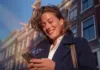 In Spagna raddoppiano i risparmiatori ma cala il risparmio medio per persona Una donna sorridente guarda il proprio smartphone mentre si trova all’aperto, con edifici sullo sfondo.