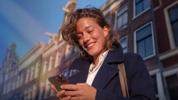Una donna sorridente guarda il proprio smartphone mentre si trova all’aperto, con edifici sullo sfondo.