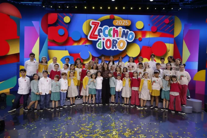 Un gruppo di bambini e adulti posa su un palco colorato; sullo sfondo compare la scritta «Zecchino d’Oro 2025».