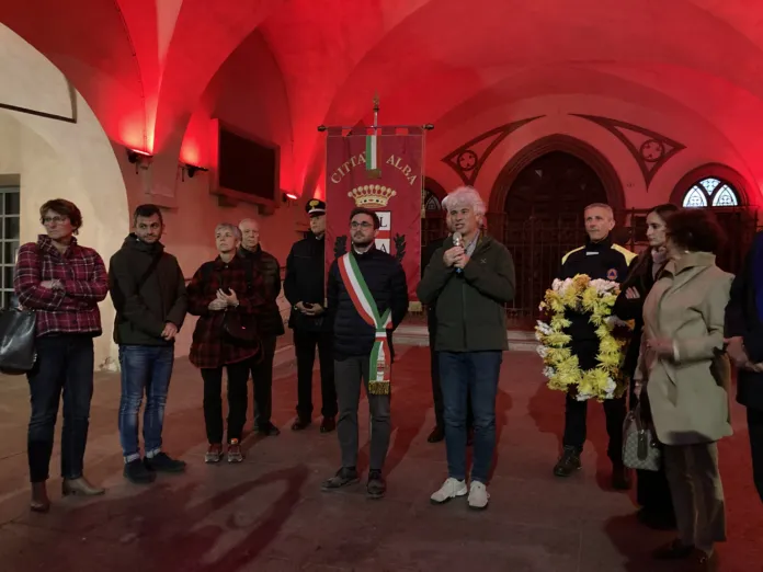 Un gruppo di persone è riunito in un interno illuminato di rosso; al centro si vede uno stendardo con lo stemma di Alba.
