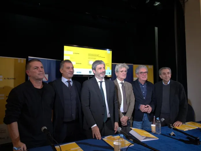 Sei persone in piedi dietro un tavolo; sullo sfondo si vedono pannelli e uno schermo.