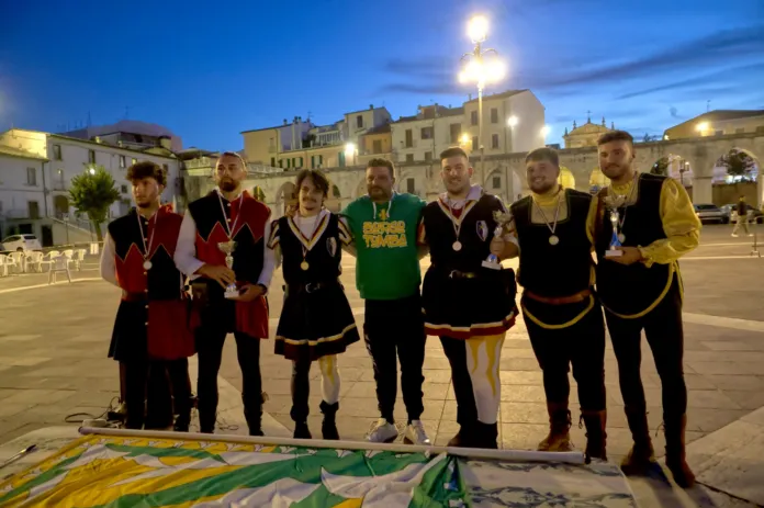 Un gruppo di persone in abiti storici è in posa in una piazza, dietro una bandiera distesa a terra.
