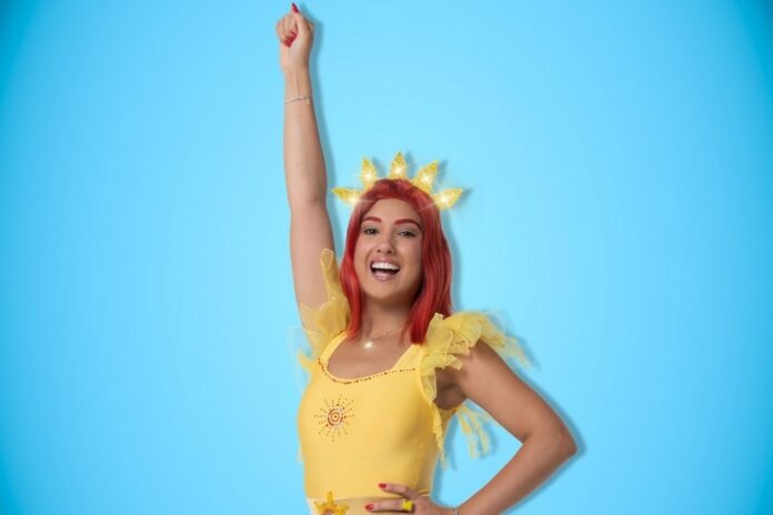 Lucilla in abito giallo con corona a raggi di sole, posa sorridente su sfondo azzurro chiaro.