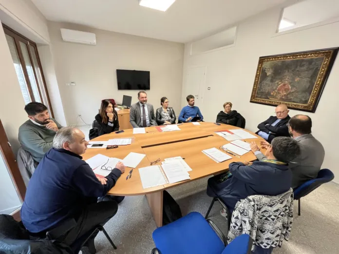 Persone sedute attorno a un tavolo con documenti e fogli; nella stanza si vedono una finestra e un quadro.