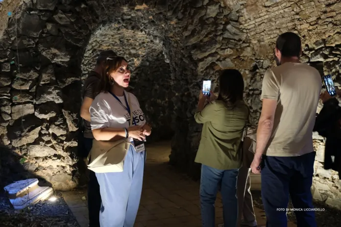Una donna parla in un ambiente sotterraneo in muratura mentre due persone la ascoltano e fotografano la visita.