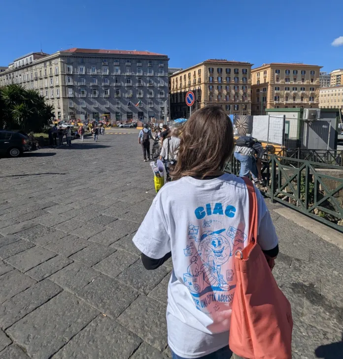 Persona vista di spalle in una piazza, con una maglietta recante la scritta CiAc e una borsa a tracolla.