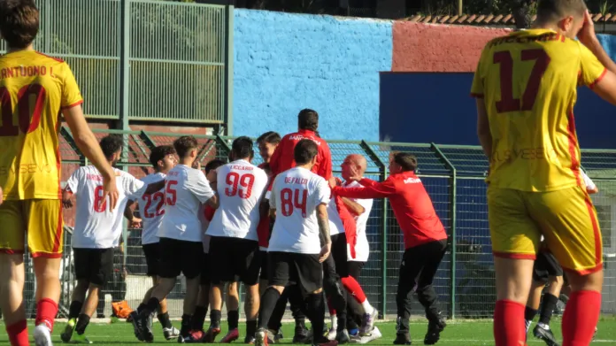 Immagine di archivio di una partita di calcio tra due squadre, con alcuni giocatori in maglietta gialla e altri in maglietta bianca con numeri rossi, in un campo all'aperto.