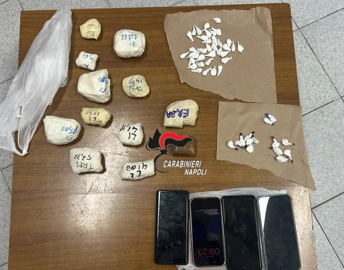 Sostanze stupefacenti e telefoni cellulari disposti su un tavolo con il simbolo dei Carabinieri di Napoli.