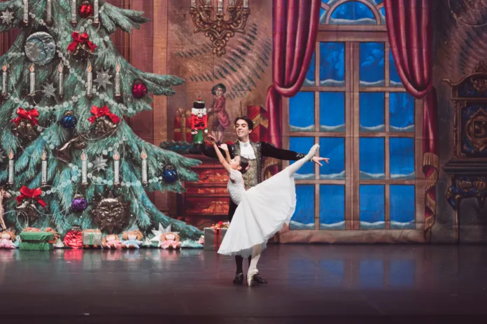 Due danzatori in costume si esibiscono accanto a un albero di Natale decorato, su un palcoscenico con una finestra sullo sfondo.
