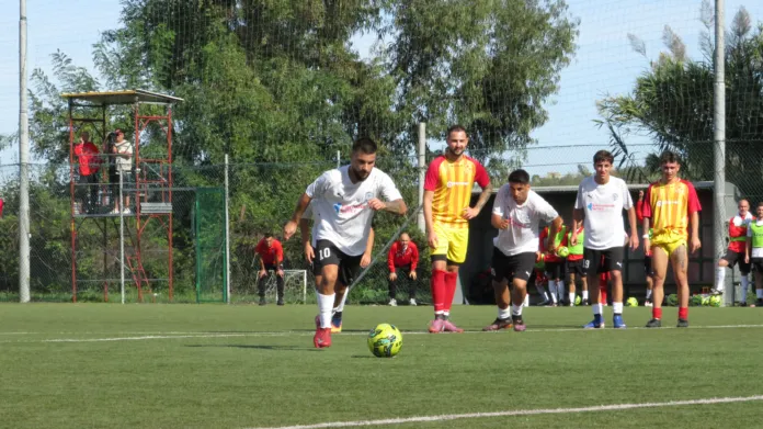 Un calciatore in maglia bianca si avvicina al pallone; altri giocatori sono disposti dietro di lui su un campo da calcio.