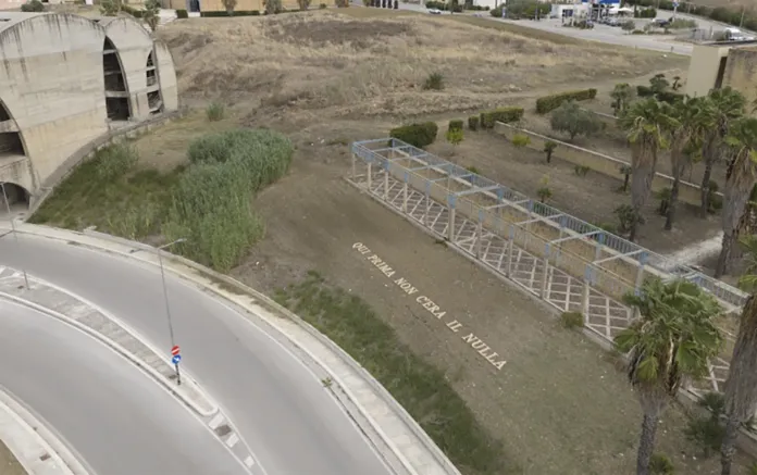 Veduta dall’alto di un’area verde con la frase «Qui prima non c’era il nulla» disposta sul terreno, accanto a una strada e a una struttura lineare.
