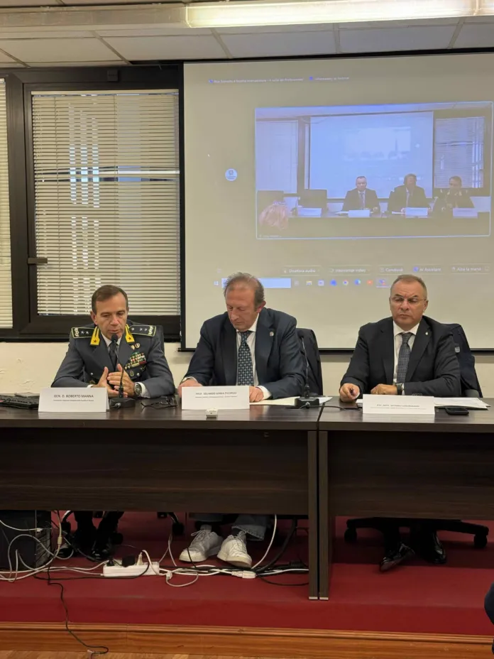 Tre uomini siedono a un tavolo con microfoni e targhette; alle loro spalle è proiettata una videoconferenza.
