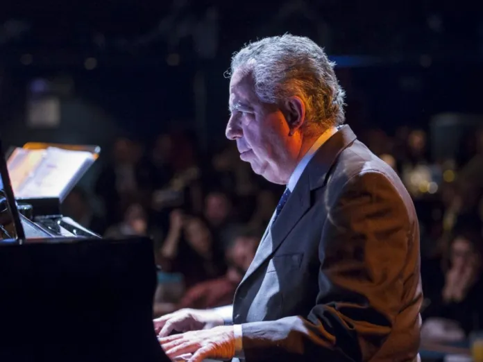 Un uomo suona il pianoforte davanti a un pubblico, in una sala poco illuminata.