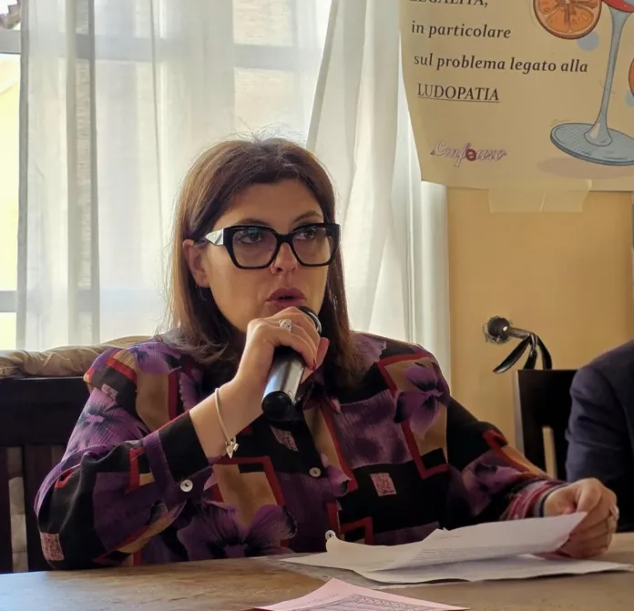 Una donna con gli occhiali tiene un microfono e alcuni fogli, seduta a un tavolo.