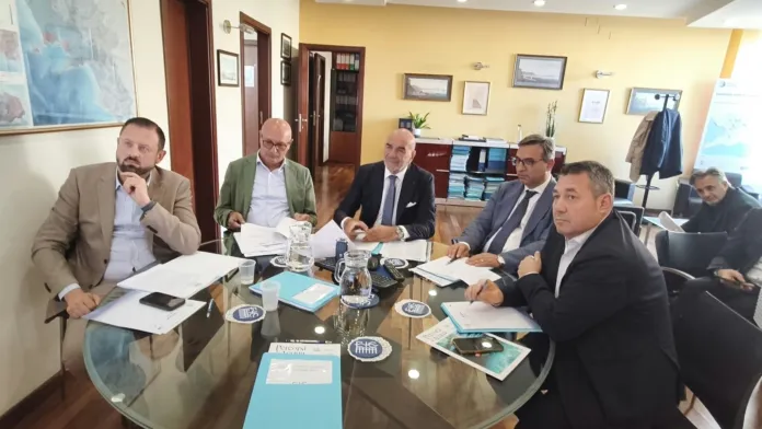 Persone sedute attorno a un tavolo di vetro in una sala riunioni, con documenti e bicchieri sul tavolo.