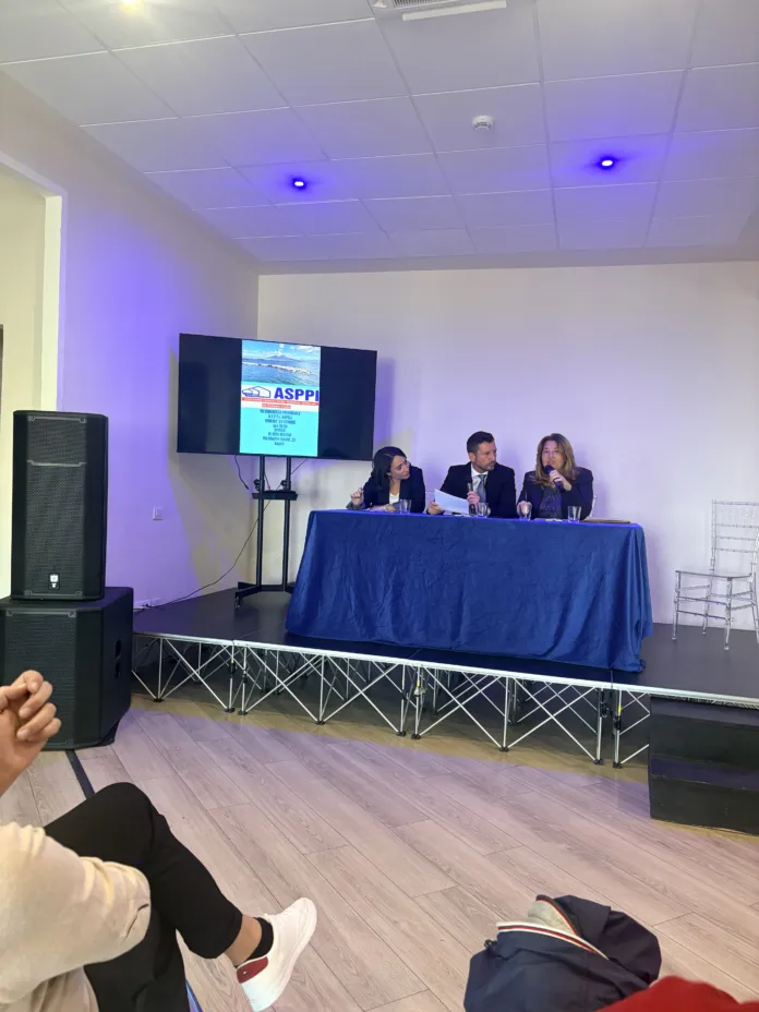 Foto di un evento congressuale con relatori seduti a un tavolo e schermi con presentazioni, scena di un congresso.