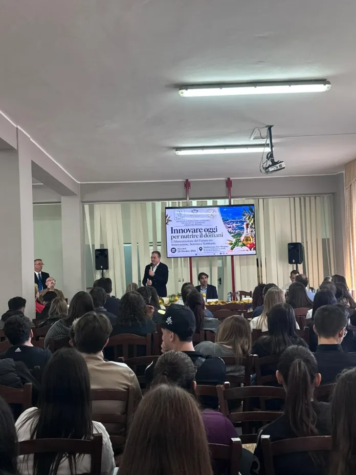 Una persona parla al microfono davanti a un tavolo con altri partecipanti; sullo schermo è visibile il titolo “Innovare oggi per nutrire il domani”.