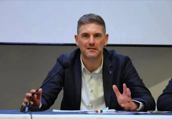 Uomo seduto a un tavolo con microfono e documenti davanti, mentre parla.