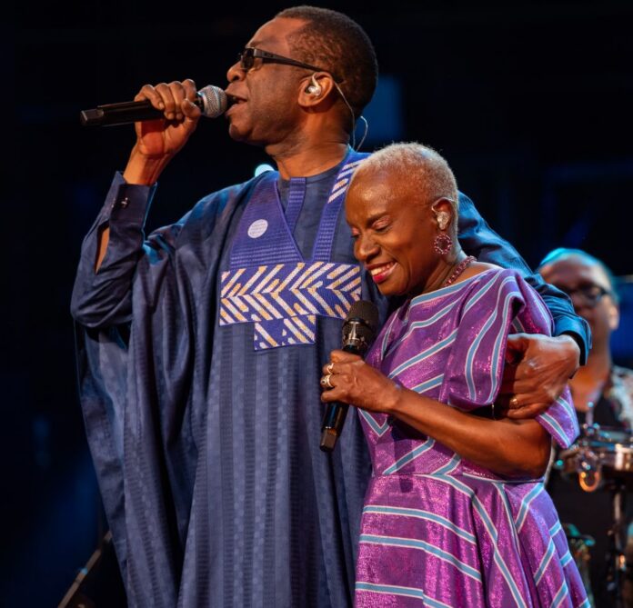 ANGÉLIQUE KIDJO e YOUSSOU N’DOUR insieme sul palco durante un concerto, entrambi con microfono in mano e abbigliamento colorato.