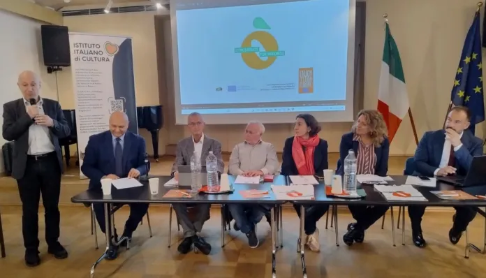 Un uomo parla al microfono accanto a un tavolo con sei persone sedute; sullo sfondo si vedono uno schermo e le bandiere italiana ed europea.
