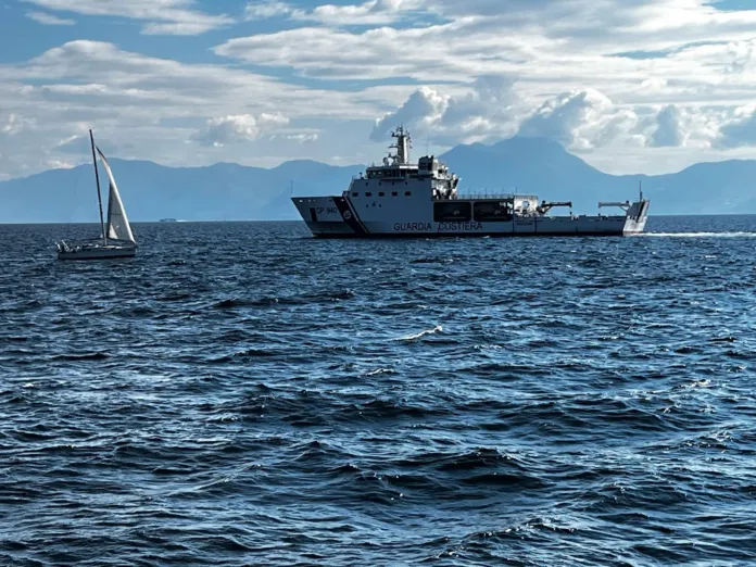 Una barca a vela in mare accanto a una nave con la scritta «Guardia Costiera».