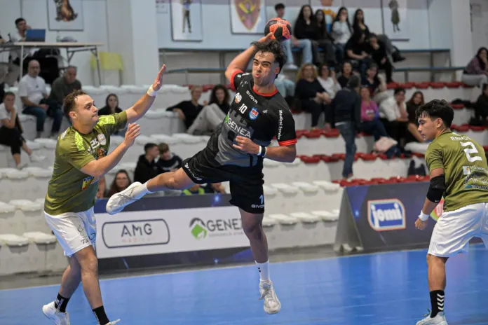Un giocatore in maglia scura salta con il pallone mentre due avversari in maglia verde cercano di contrastarlo; sullo sfondo si vedono il pubblico e le tribune.