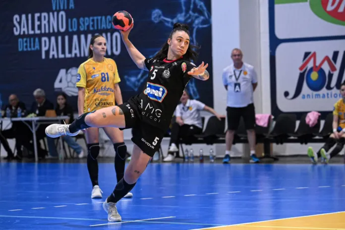 Una giocatrice in maglia nera salta e lancia il pallone, mentre un’avversaria in maglia gialla la segue sul campo.
