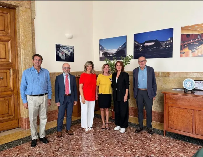 Sei persone in piedi in una sala con pareti chiare e pavimento in marmo; alle loro spalle sono appese alcune fotografie.