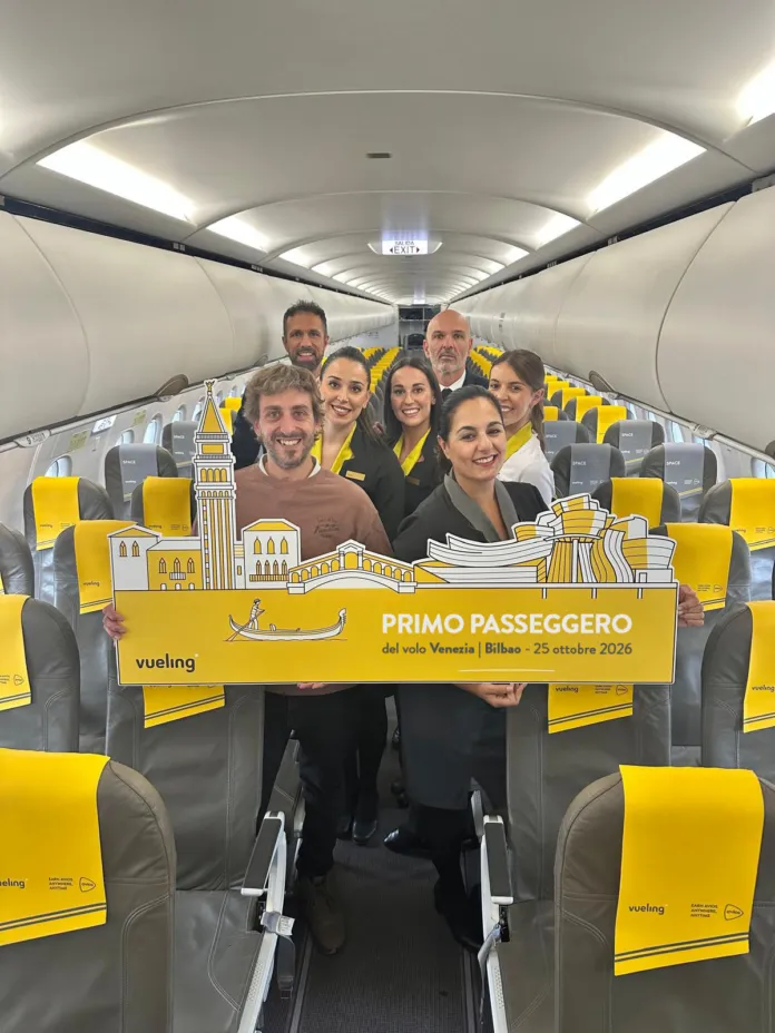 Un gruppo di persone è in piedi nel corridoio di un aereo, tra sedili gialli e grigi, e tiene un cartello con la scritta «Primo passeggero» e i nomi Venezia e Bilbao.
