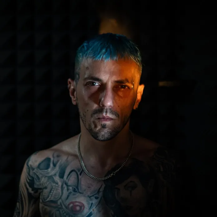 Primo piano di un uomo con capelli azzurri e tatuaggi, su sfondo scuro.