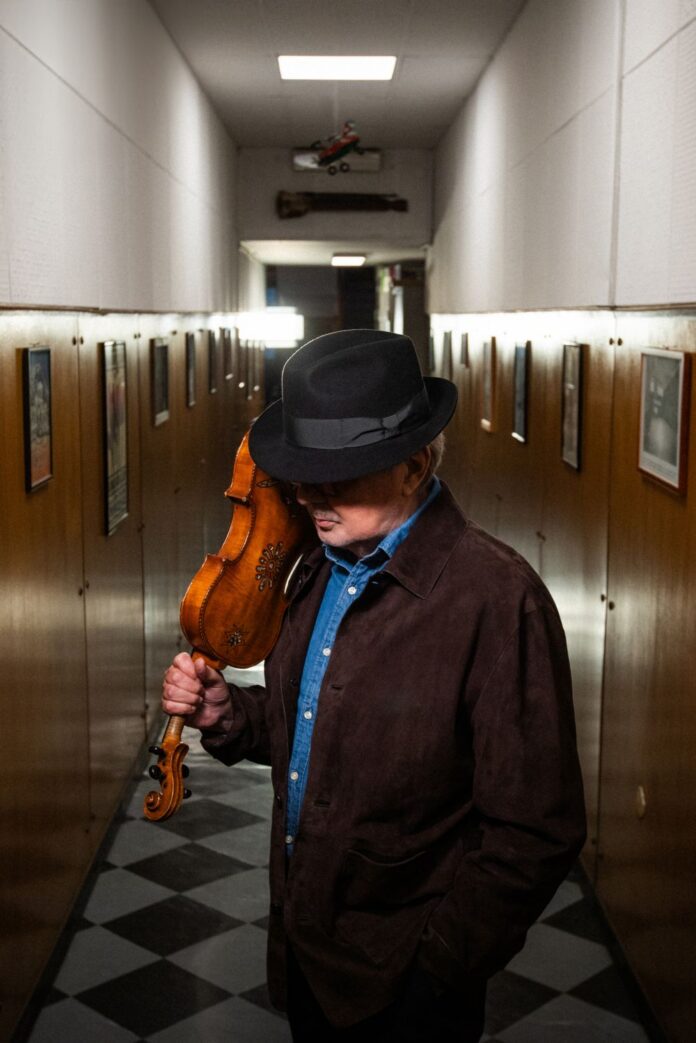 Mauro Pagani con cappello e giacca scura che tiene un violino su una spalla in un corridoio lungo con pareti e quadrotti alle pareti.