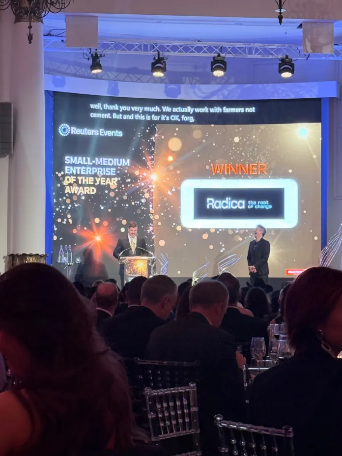 Sala con pubblico e palco; sullo schermo si leggono “Reuters Events”, “Winner” e “Radica”.