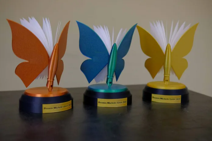 Tre trofei a forma di farfalla, con targhette del Premio Michele Serio 2026, appoggiati su un tavolo.