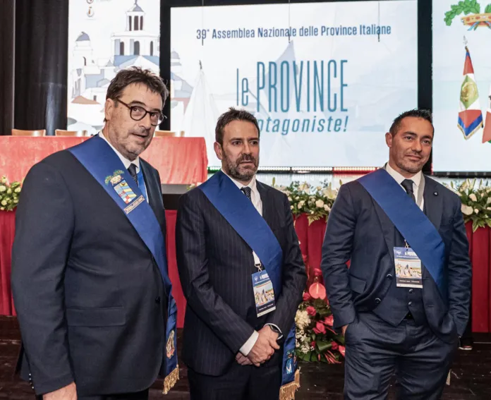 Tre uomini in abito scuro con fasce blu; sullo sfondo, uno striscione con la scritta «39ª Assemblea Nazionale delle Province Italiane».