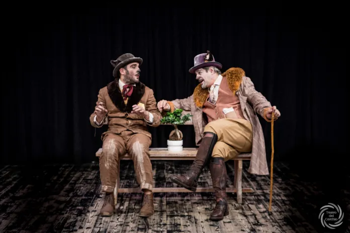 Due uomini in costume di scena sono seduti su una panchina, su un palco con tende nere sullo sfondo.