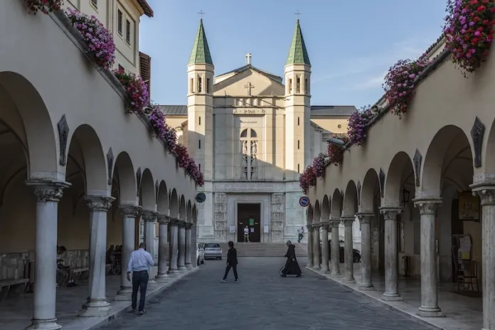 Un porticato fiancheggia una piazza; sul fondo si vede una chiesa con due torri campanarie.