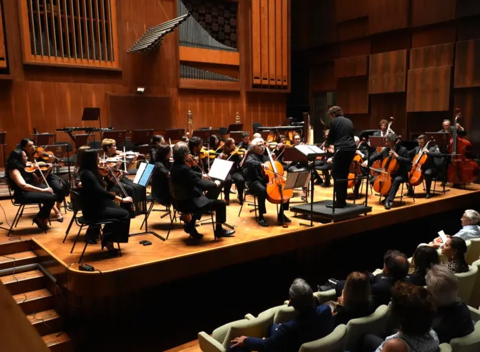 Orchestra e direttore su un palco in una sala da concerto, con parte del pubblico in primo piano.