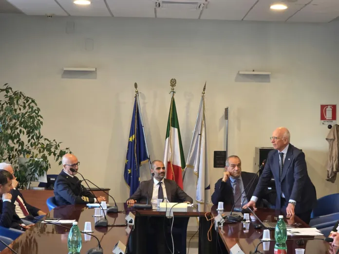 Persone sedute attorno a un tavolo in una sala con bandiere sullo sfondo; un uomo è in piedi accanto al tavolo.
