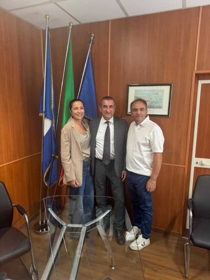 Tre persone in piedi in un ufficio con bandiere e pareti in legno, foto illustrativa dell'incontro istituzionale sulle bonifiche.