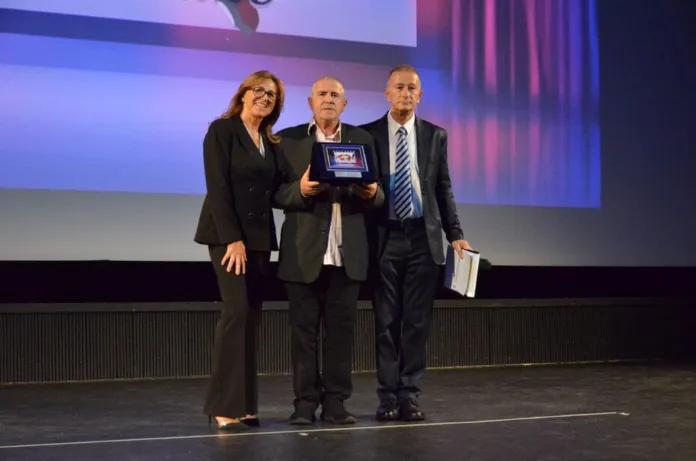 Immagine di una cerimonia di premiazione con tre persone sul palco, una delle quali tiene un premio in mano, in un teatro.