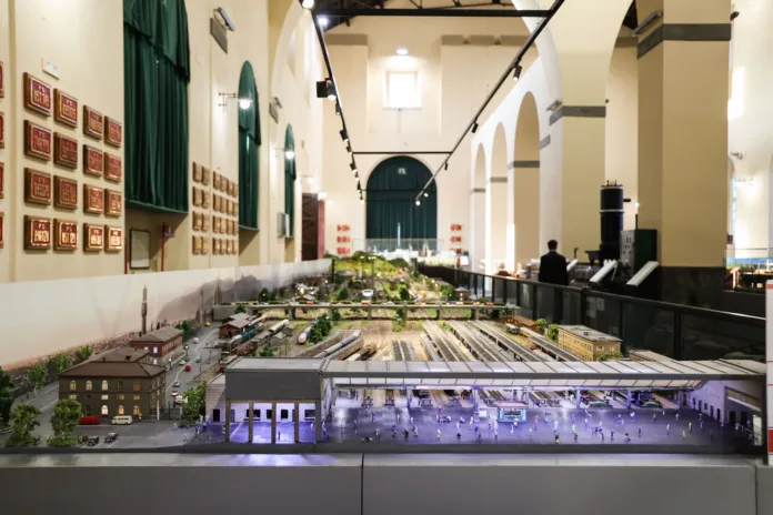 Un plastico ferroviario con binari, edifici e paesaggi in miniatura, esposto in una sala.