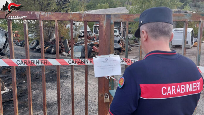 Area recintata con nastri dei Carabinieri e rifiuti visibili all'interno, con un carabiniere di spalle davanti al cancello.