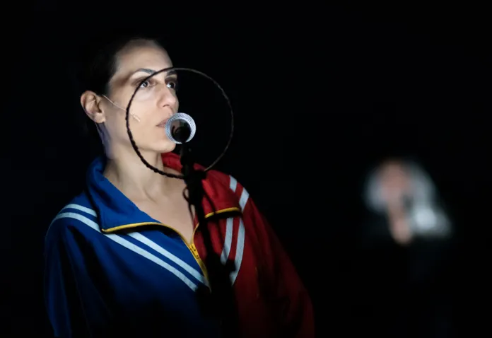 Scena teatrale con un'attrice in primo piano con costume colorato e microfono circondato da un ring nero, sfondo scuro.