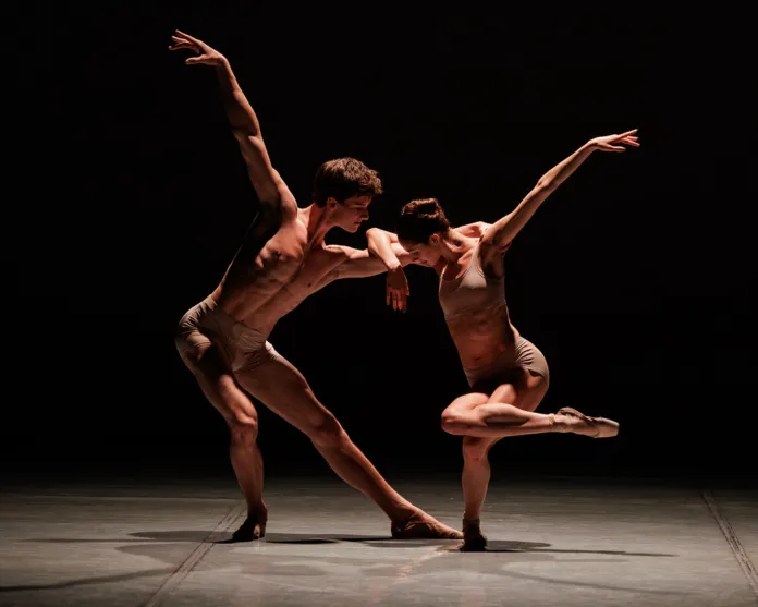 Fotografia di scena di due ballerini in una performance di danza su un palco oscuro, con luci focalizzate su di loro.